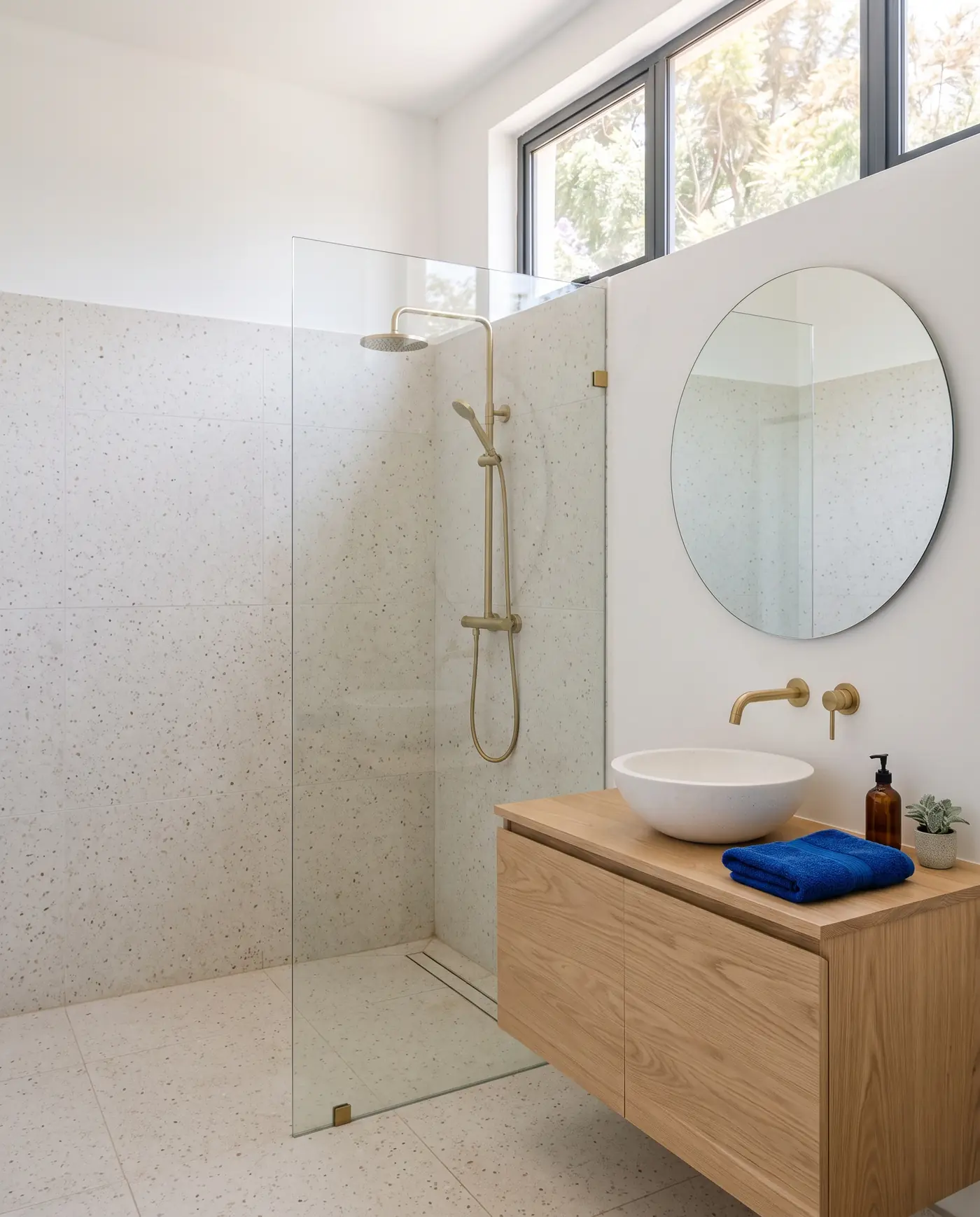 A bathroom with a walk-in shower behind a glass panel, terrazzo floors and an oak vanity